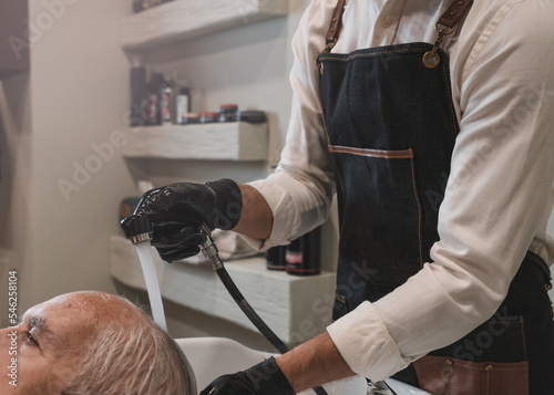 Peluquero irreconocible lavando el pelo a cliente senior, con grifo de agua, en la peluquería.