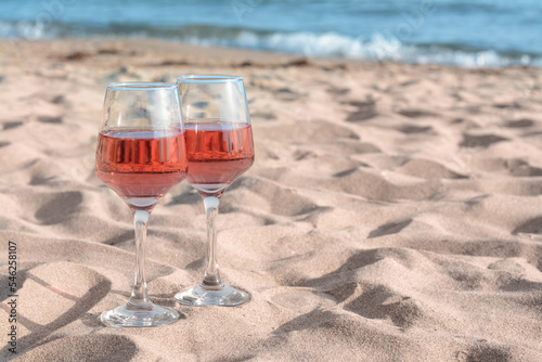 Glasses of tasty rose wine on sand near sea, space for text