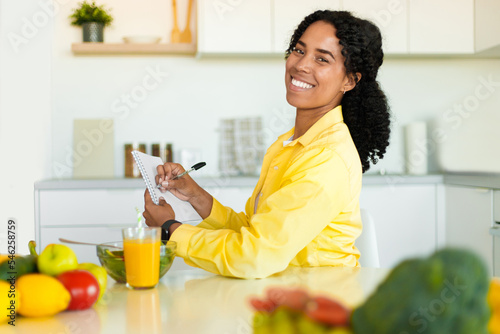 Happy fit african american woman writing daily ration diet or menu in notebook, sitting at kitchen table, copy space