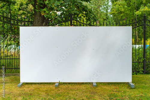 A template with a blank white sign next to a fence made of metal bars. Ground large billboard in the grass near the fence of the park.