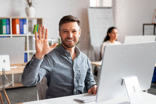 Smiling attractive millennial caucasian man manager working at computer, waving hand at camera