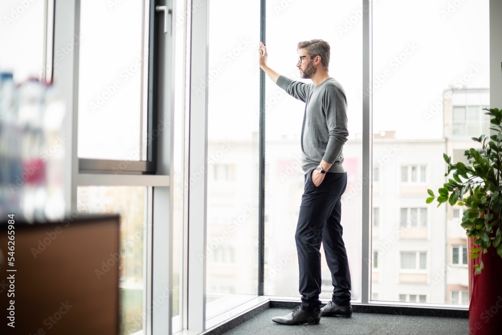 Shot of thinking businesman standing at the office and looking out of city Shot of thinking businesman standing at the office and looking out of city