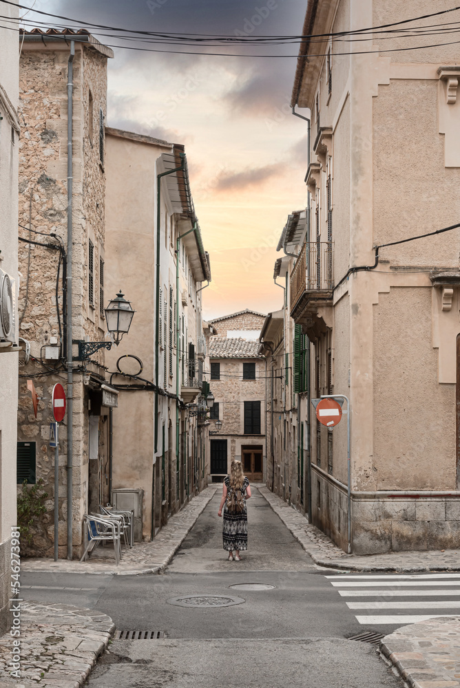rear view of woman in long dress standing in middle of narrow street amidst historic houses in old spanish town of Soller