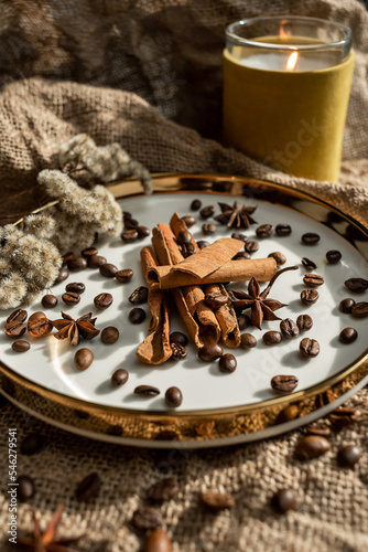 An elegant composition of candles, plates with a golden border, on which spices - anise, cinnamon and coffee beans lie. Against the background of burlap