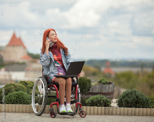Fotografie Beautiful red-haired girl on a wheelchair in a city park with a smartphone and a laptop