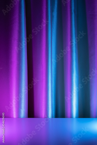 Silky shiny curtain with folds in neon blue and purple light behind the reflective table with space for product