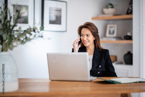 Happy middle aged woman sitting at table and using laptop while working from home