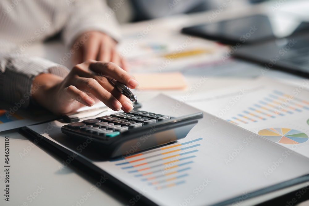 Businessman is using a calculator to calculate company financial figures from earnings papers, a businessman sitting in his office where the company financial chart is placed. Businessman is using a calculator to calculate company financial figures from earnings papers, a businessman sitting in his office where the company financial chart is placed.