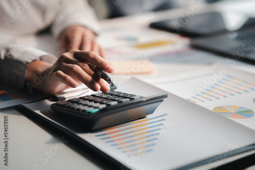 Businessman is using a calculator to calculate company financial figures from earnings papers, a businessman sitting in his office where the company financial chart is placed.