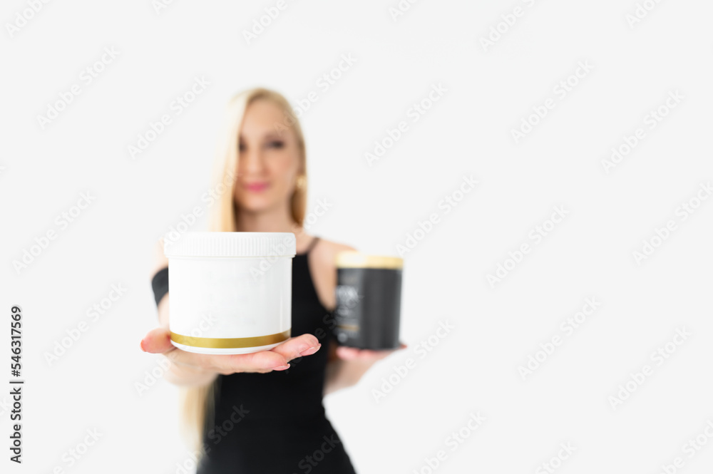 a blonde on a white background with long beautiful hair in a black dress in the background holds a white plastic jar with a gold border and a black one in the other outstretched in her hand. Care a blonde on a white background with long beautiful hair in a black dress in the background holds a white plastic jar with a gold border and a black one in the other outstretched in her hand. Care