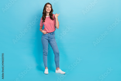 Full length photo of adorable pretty girl dressed red t-shirt pointing thumb looking empty space isolated blue color background