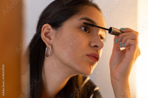 Young ethnic lady looking in mirror while applying mascara