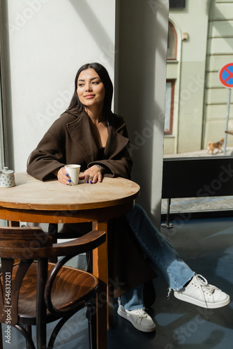 full length of brunette woman in trendy autumn outfit sitting in cafe in prague. photo