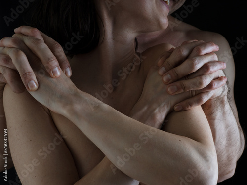 A man hugs a woman from behind. Naked bodies. Plexus of the hands. Close-up photo without a face on a dark background.