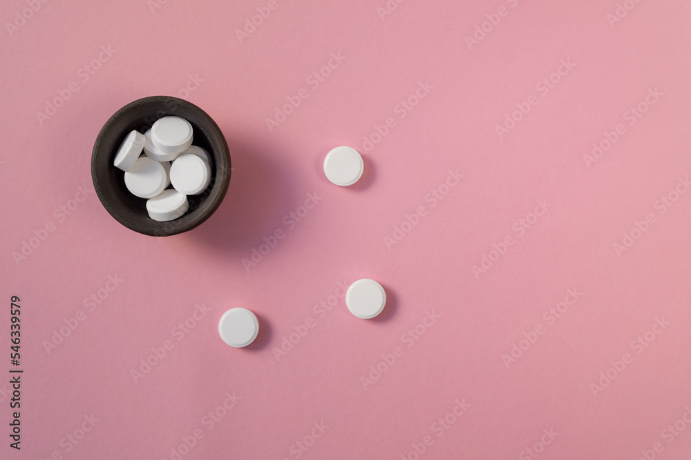 White large vitamin tablets in a dark ceramic bowl on a pink background. Medicine and health.