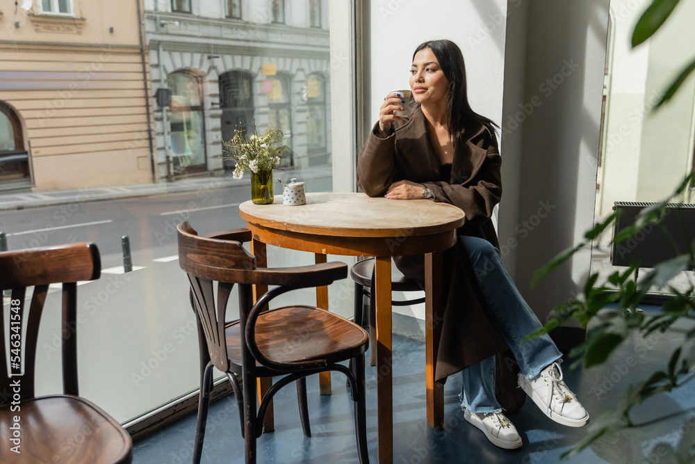 full length of brunette woman looking at city street while sitting in cafe in prague. full length of brunette woman looking at city street while sitting in cafe in prague.