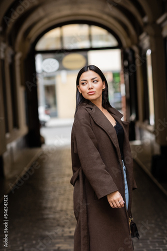 pretty brunette woman in trendy coat looking away near blurred arch building in prague. photo