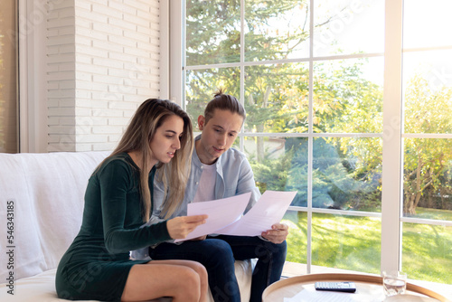 Worried young couple reading documents about mortgage loan insurance contract terms sitting in the living room with the garden in the background