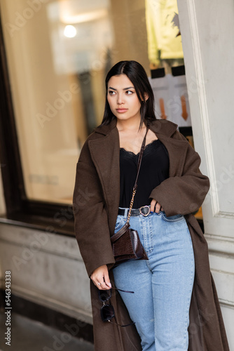 pretty woman in trendy coat and jeans looking away near blurred building on street in prague. photo