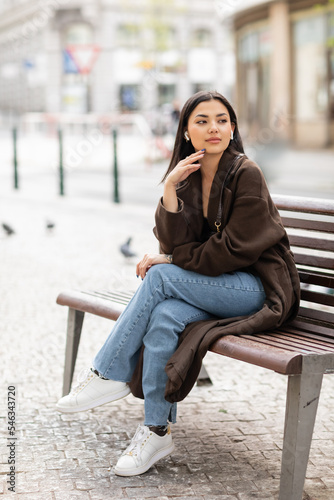 pretty woman in trendy autumn clothes listening music in wireless earphones on bench in prague and looking away. photo