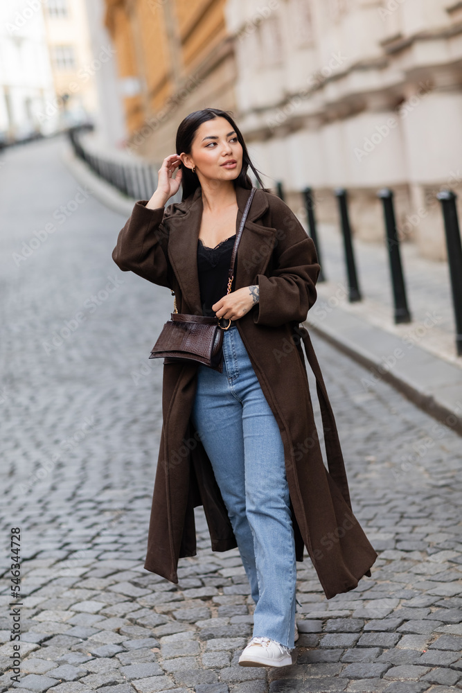 brunette woman in trendy coat with crossbody fixing hair and looking away on blurred street in prague.