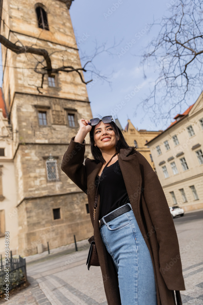 Low angle view of happy traveler in coat holding sunglasses near blurred buildings in Prague.