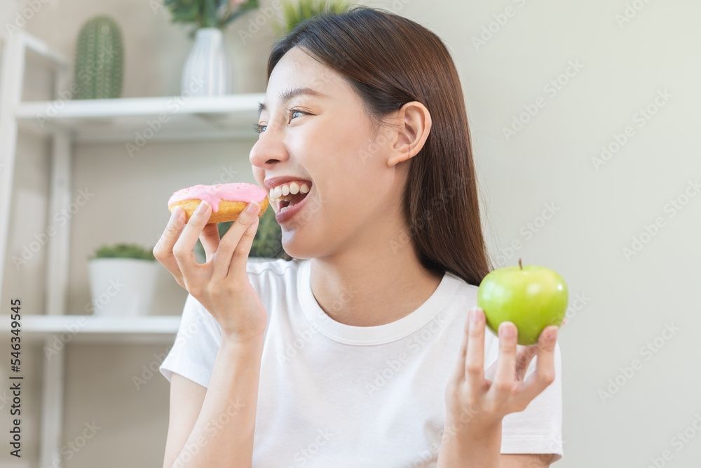 Diet, Dieting. Close up hand of asian young woman confused with food temptation choose, choice pink sweet donut or green apple at home, eat food for good healthy, health when hungry, weight loss. Diet, Dieting. Close up hand of asian young woman confused with food temptation choose, choice pink sweet donut or green apple at home, eat food for good healthy, health when hungry, weight loss.