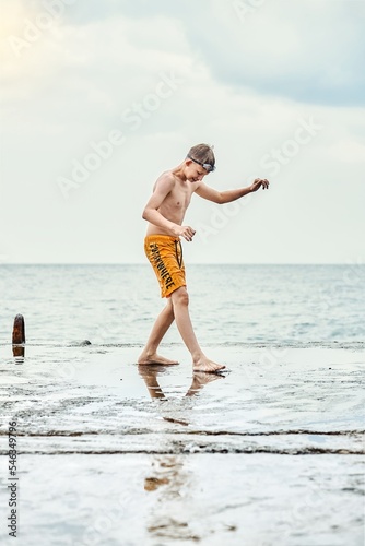 Teenager wearing yellow trunks stands on sea pier on dull day. Teen schoolboy tries to keep his balance on a wet slippery concrete pier against seascape