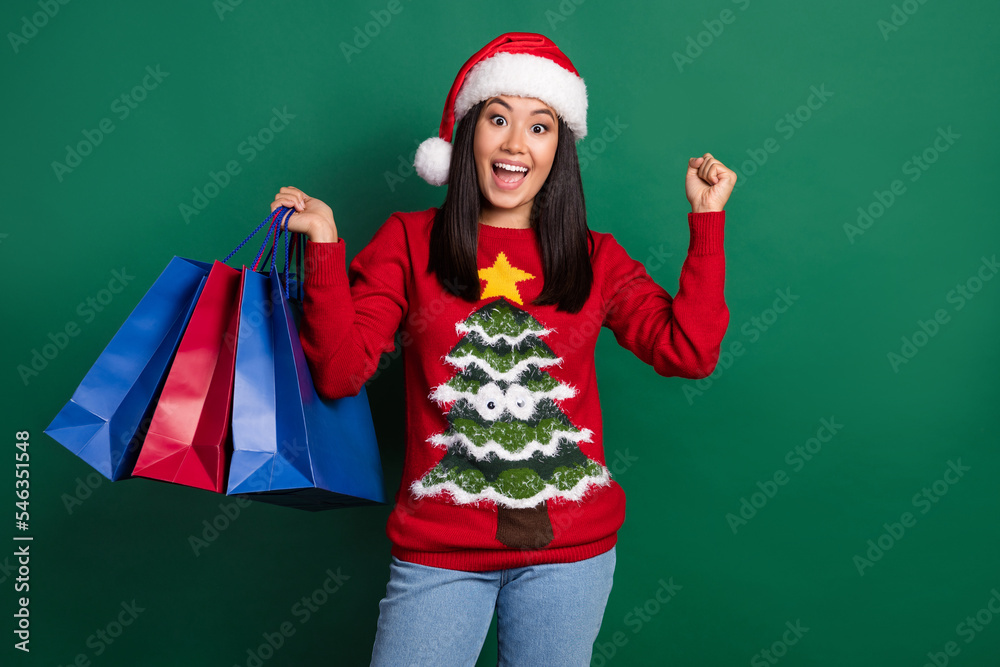 Photo of funky japanese woman wear red old ugly sweater raise hand up hold many bargains from boutique xmas sale isolated on green color background Photo of funky japanese woman wear red old ugly sweater raise hand up hold many bargains from boutique xmas sale isolated on green color background