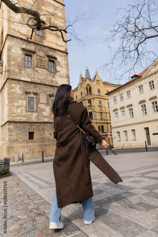 Young woman in coat holding sunglasses while walking on urban street in Prague.