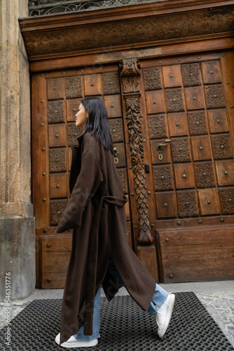 side view of brunette woman in stylish coat walking near carved wooden door on street in prague. photo