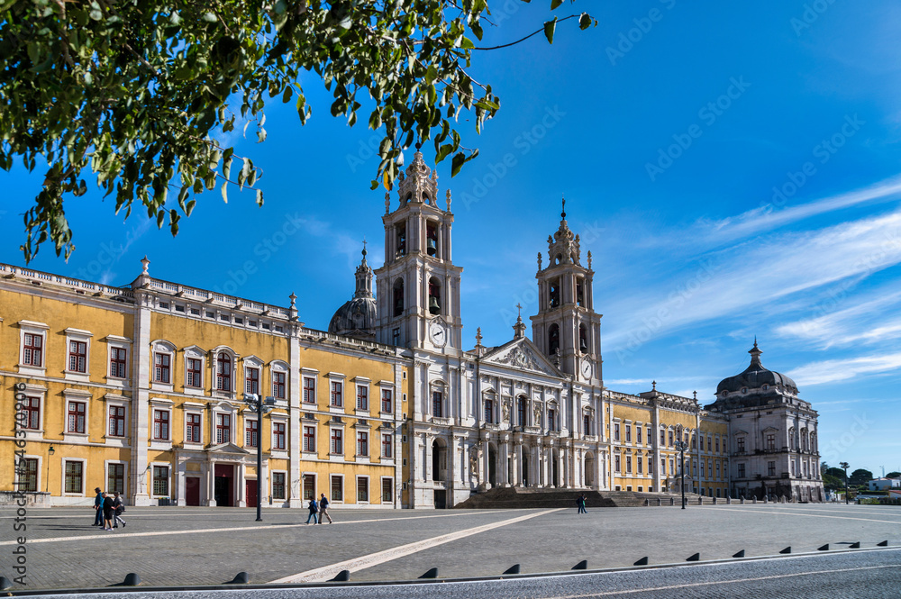 Obraz premium The grand Mafra National Palace in Portugal, with its impressive yellow and white facade and prominent bell towers, under a clear blue sky.
