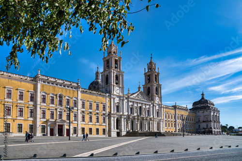 The grand Mafra National Palace in Portugal, with its impressive yellow and white facade and prominent bell towers, under a clear blue sky.