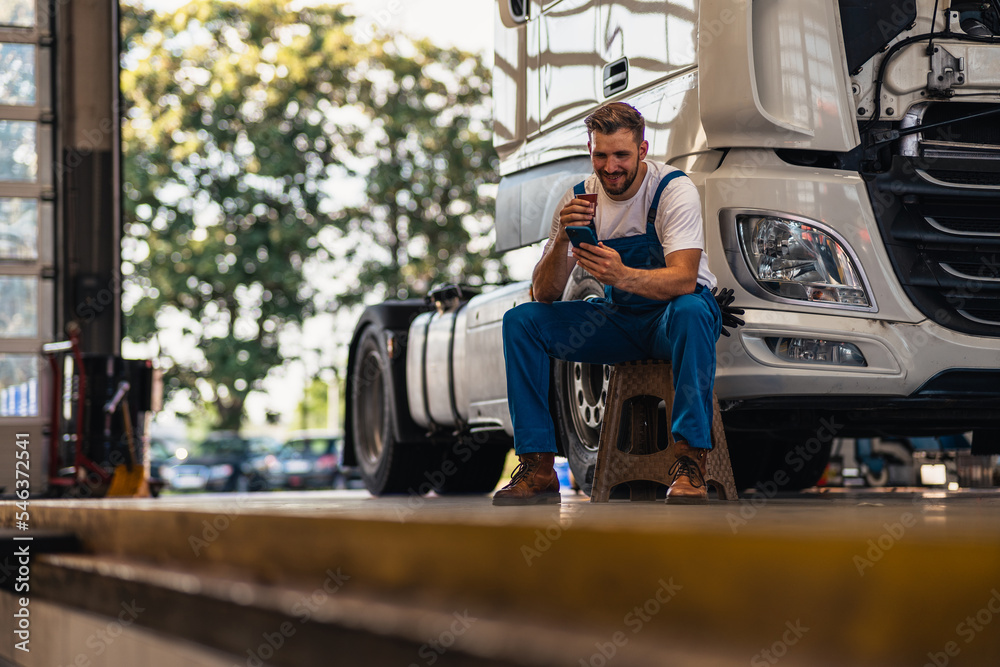 Naklejka premium A bottom view of a mechanic drinking tea or coffee from a cup and sitting leaning against a truck in a workshop. A young caucasian men mechanic. Truck service, repair. Rest and break at work.
