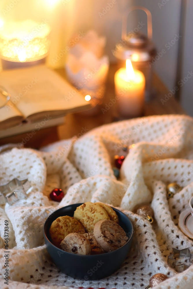 Cups of tea or coffee, bowl of cookies, soft blanket, lit candles, books and various Christmas decorations on the table. Selective focus.