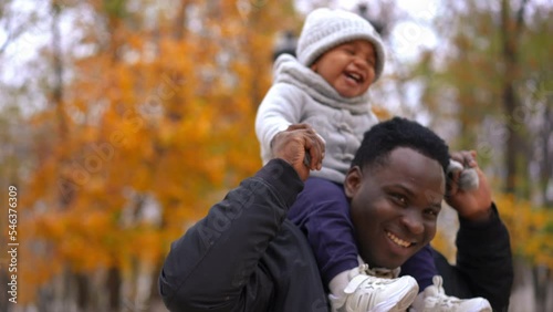 Portrait of happy young man spinning with toddler boy on shoulders smiling. African American father resting with son in autumn park enjoying overcast day outdoors