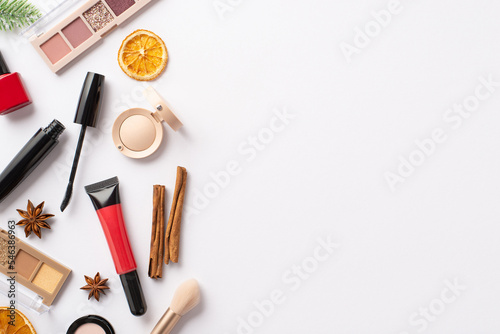 Top view photo of decorative cosmetics red lip gloss nail polish mascara eyeshadow palettes brush fir branch dried orange slices cinnamon sticks on isolated white background with copyspace
