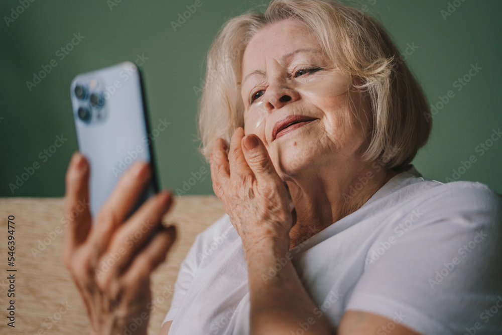 Joyful senior woman with collagen patches under her eyes sitting with her smartphone on the sofa in the morning. Contemporary portrait. Joyful senior woman with collagen patches under her eyes sitting with her smartphone on the sofa in the morning. Contemporary portrait.