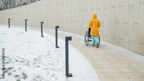 Caucasian woman carries an empty wheelchair through the park in winter.