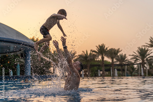 Dad and son are playing in the swimming pool. The father tosses the son up.