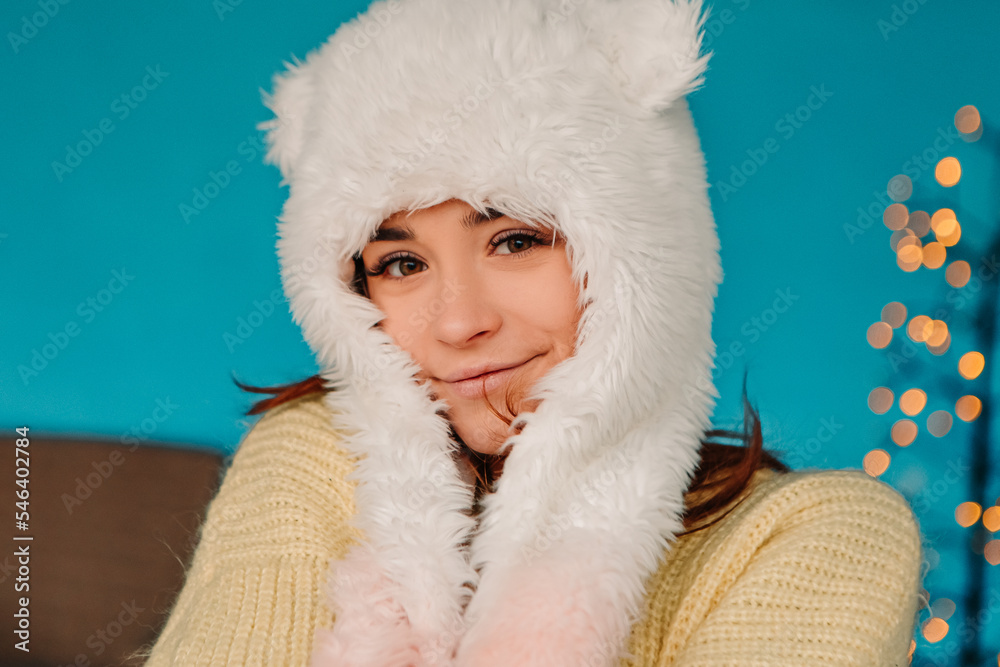 Young pretty smiling woman celebrating dressed warm sweater and woolen hat, standing posing. Winter market holidays fair, travel spirit resort. New Year Christmas decorations, garlands
