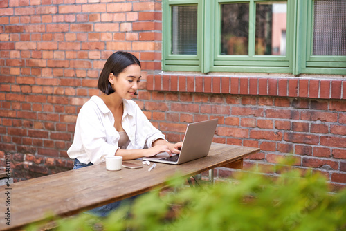 Stylish young urban girl in outdoor cafe, sitting on bench with laptop, smiling, browing on computer photo