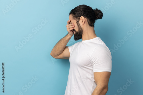 Don't want to look at this. Side view of scared man with beard wearing white T-shirt standing, covering eyes with hand, refusing to watch. Indoor studio shot isolated on blue background.