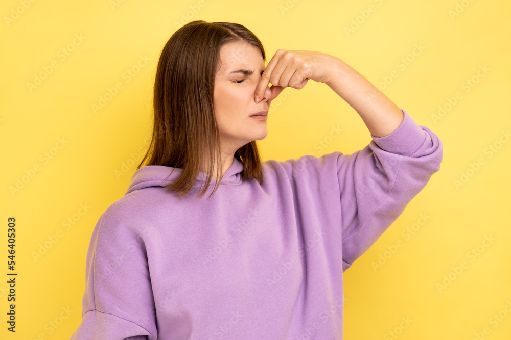 Portrait of dark haired woman grimacing with disgust, holding breath, pinching nose with fingers to avoid bad smell, awful odor, wearing purple hoodie. Indoor studio shot isolated on yellow background Portrait of dark haired woman grimacing with disgust, holding breath, pinching nose with fingers to avoid bad smell, awful odor, wearing purple hoodie. Indoor studio shot isolated on yellow background