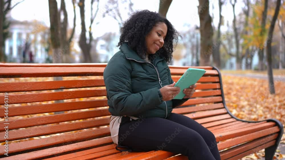Beautiful smiling young woman surfing Internet sitting on bench in autumn park. Portrait of positive confident African American lady messaging online outdoors Beautiful smiling young woman surfing Internet sitting on bench in autumn park. Portrait of positive confident African American lady messaging online outdoors