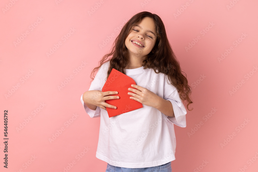 Portrait of satisfied little girl wearing white T-shirt standing open red envelope with congratulations, reading romantic letter, being touched. Indoor studio shot isolated on pink background. Portrait of satisfied little girl wearing white T-shirt standing open red envelope with congratulations, reading romantic letter, being touched. Indoor studio shot isolated on pink background.