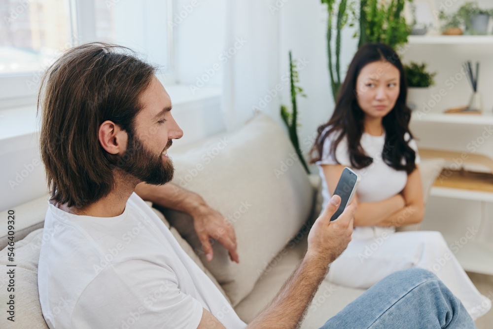 Fototapeta premium A man looks at his phone screen and smiles during an argument with his girlfriend. The angry and hurt woman looks in his direction and is sad. Family discord at home, phone addiction