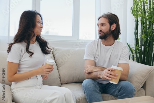 Man and woman sitting at home on the couch in white stylish t-shirts drinking coffee out of crab cups from a coffee shop and having fun chatting smiles and laughter at home. Male and female friendship