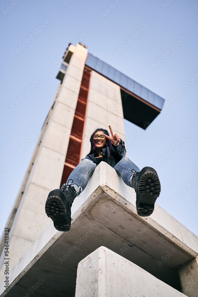low angle shot of brunette latina girl in big black boots sitting on the corner of the building smiling at camera next to the concrete building low angle shot of brunette latina girl in big black boots sitting on the corner of the building smiling at camera next to the concrete building