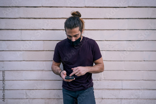 caucasian young man in black t-shirt and face mask looking and touching his smartphone screen near the wall